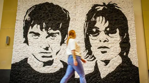 A black and white mural of noel and liam gallagher, both looking sternly forward. They have short cropped hair and liam is wearing round sunglasses. The texture of the bucket hats can be seen. the mural is up against a yellow wall. there is a blonde pony tailed woman in blue jeans and a white top walking right in front of the mural and her legs and arms are blurred by motion