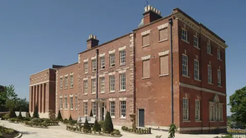 A large three-story rectangular red brick historic builidng, there are small cone shaped tree in the grounds at the front of the building