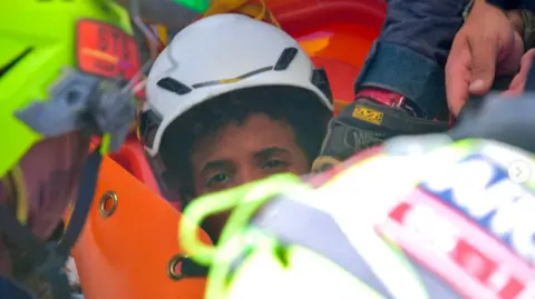 ungrd_oficial the picture shows half of the boys face - the rest is obscured by rescue workers. He's wearing a white hard hat and has dark curly hair.