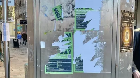 Michael Enea Posters torn off a telephone box along a high street road in a shopping district, with people walking across.