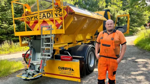 Road worker in orange high-visibility clothing standing beside a yellow gritting truck marked Spreading on a rural roadside with trees in the background.