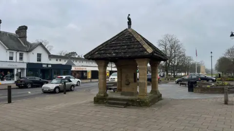 BBC An image of the centre of Cranleigh, including a former drinking fountain, which has become a well known monument in the village. Cars drive past on the high street. Shops, including Sainsbury's, are in the background. Also in the background is the local war memorial.