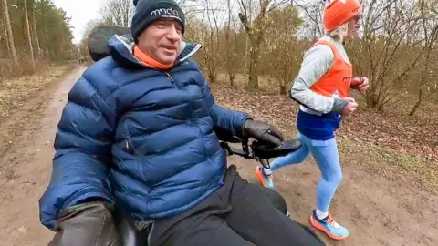 A man in a powerchair, dressed in warm clothes, drives down a country track alongside a jogging woman