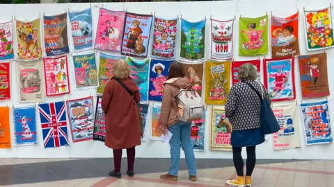 Visitors looking at a large exhibition of brightly coloured tea towel artworks displayed on lines inside a gallery space.