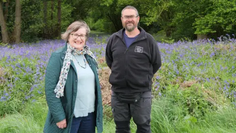Rachael Murray An outdoor daylight photo of two middle-aged people standing in front of a large patch of bluebells in front of an area of woodland. On the left is a smiling woman with shirt, dark, greying hair and glasses, wearing a green coat and a pale green top, blue trousers and a floral scarf. On the right is a man with short, dark greying hair and a beard, also smiling and wearing glasses, wearing a black top and charcoal-coloured trousers.