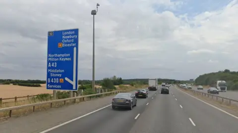 Google Junction 10 of the M40, a three-lane road. Cars can be seen driving along it. On the left, a blue sign shows an exit to the A43.