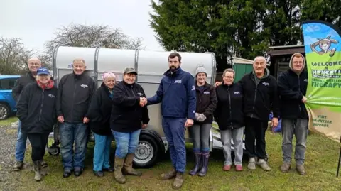 Hensell Goat Rescue Care Farm A group of people wearing branded fleeces, jeans and boots stood on a farm in front of a trailer. 