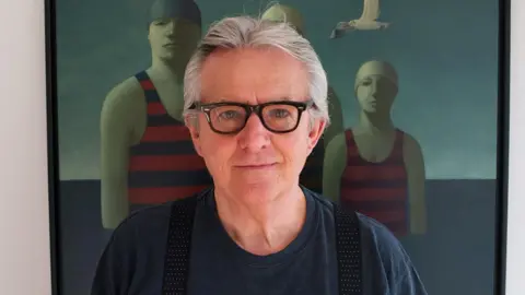 George Underwood George Underwood wearing black frame glasses and and a blue top, standing in front of a painting of three people standing in water