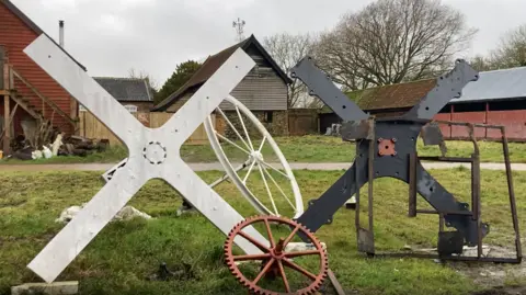 Luke Deal/BBC A large white part of a windmill and a smaller black one next to it sit outside several buildings on grass. They are in the shape of a cross. Other wheel parts sit in front and behind them.