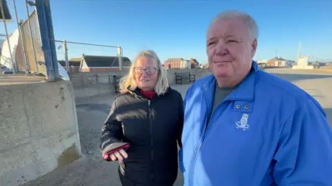 Crispin Rolfe/BBC A woman wearing a black coat with shoulder-length blonde hair and glasses and a man stood next to her with short grey hair wearing a light blue coat with an owl on it. They are stood on a promenade.