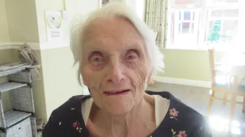 An elderly woman stood looking into the camera. She is wearing a white to with a blue jumper over it, with flowers printed on it.