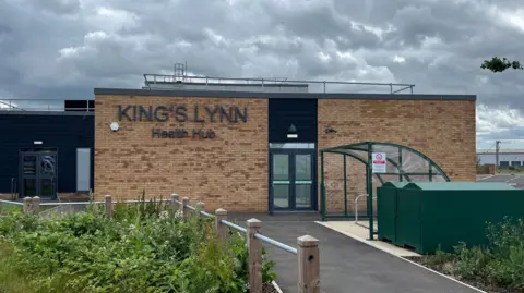 A yellow brick building with the words "King's Lynn Health Hub" written on it in dark grey letters. There is a door in the centre. In the front of the image is a bicycle shelter with a roof. There are green plants on the front left of the image. 