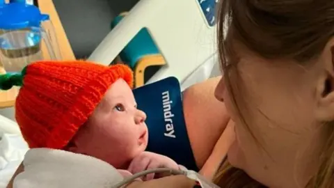 Natalie looking down at newborn baby Arthur in her arms wearing an orange hat. She has brown hair and a blood pressure monitor on her finger and is sat in a hospital bed, with a wooden table and chair beside her.