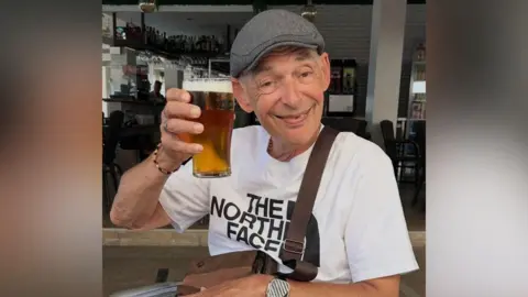 Richard Wainhouse sat at a table smiling holding a pint of beer. He is wearing a white and black tshirt and a grey flat cap with a bag on his shoulders 
