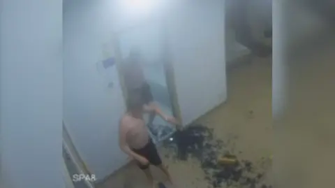 CCTV image of two boys in swimming trunks stepping out of a steam room. They have brown hair. One boy is looking at his arm and there is shattered glass on the floor around them.