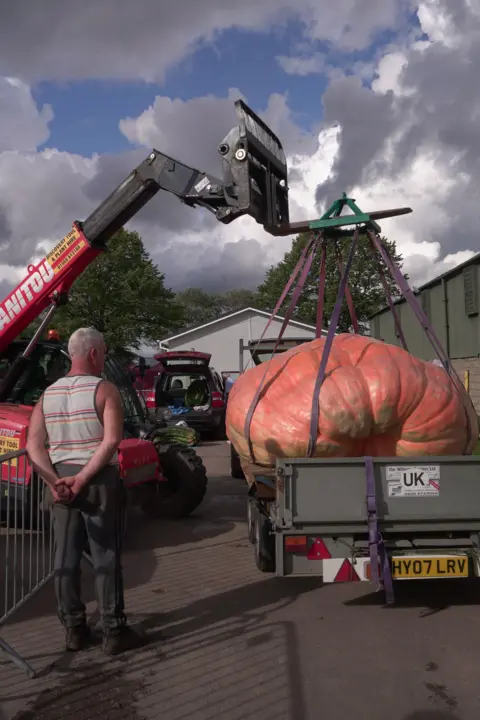 A man stands in front of a pumpkin, being lifted by a mini crane from a trailer. 