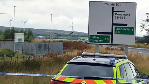 a police car is parked behind a cordon and in front of a road sign showing directions to Claudy, Dungiven, Belfast, Londonderry and Dungiven. In the backgorund is a second police car and a number of wind turbines can be seen atop hills in the distance