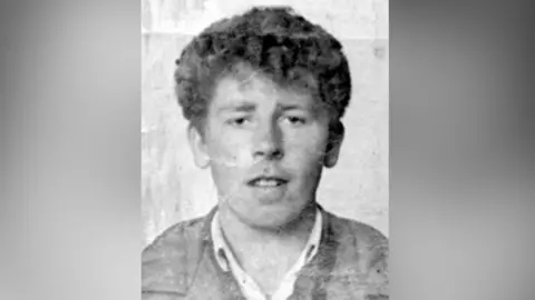 A black and white photo of Seamus Maguire - a man with short, curly hair. He is looking straight into the camera. He is wearing an open white shirt and dark jacket.