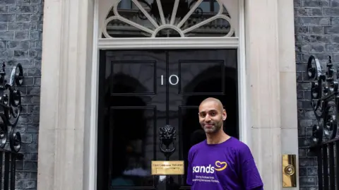 Man wearing purple Sands t shirt outside famous black number 10 door in Downing Street 