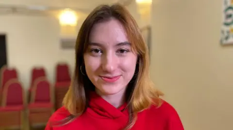 BBC Olksandra is wearing a red top and has long hair