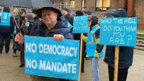 A group of people, mostly men, stand in the drizzle holding blue-coloured placards, that read NO DEMOCRACY NO MANDATE and WHO THE HELL DO YOU THINK YOU ARE?. A man stands in the foreground, holding an umbrella, looking towards the camera