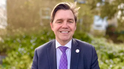 Head and shoulders image of Nik Johnson. He has brown hair swept to his right and is wearing a navy suit, blue shirt and purple tie. He is standing in front of foliage.