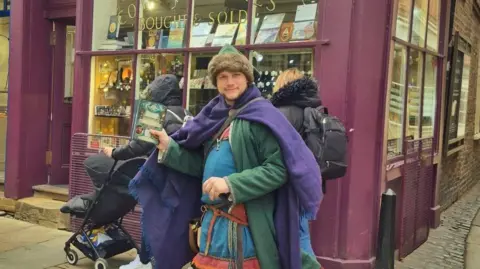 Other A man dressed in medieval‑style clothing — wearing a green coat, blue tunic, fur hat and purple cloak — stands on a cobblestone street holding a colourful booklet, with a purple‑fronted shop behind him and passers‑by in the background.