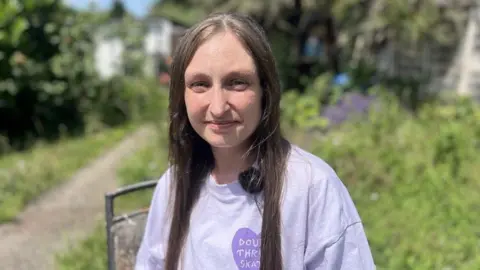 Sophie Excell wearing a purple t-shirt. She is looking at the camera and is stood outdoors, in a garden.