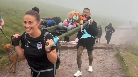 Sian Donoghue A man and Woman carrying hand weights and a bear on a stretcher up the mountain. The weather is foggy. The woman at the front is smiling off camera.