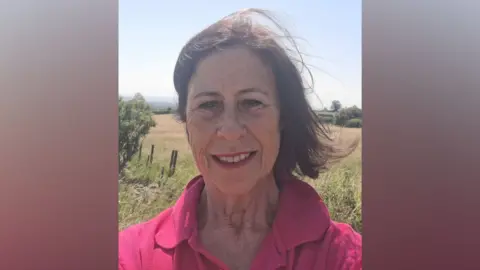 Susan Tanner Women with dark-hair, who is dressed in a pink top. She is taking a selfie stood in some green fields. There is blue skies in the background.