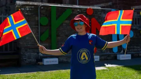 Muckle Media A boy standing holding two Orkney flags wearing a red bucket hat