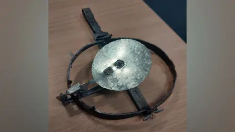 A gin trap. It comprises of a silver metal disk with a black metal ring around it. It is placed on a wooden table.