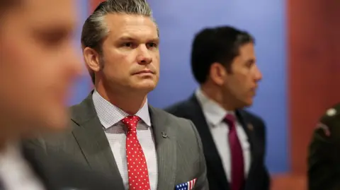 Reuters Hegseth, in blue-checked shirt with red and white polkadot tie under grey suit jacked with American flag breast pocket handkerchief, stares off to his right.