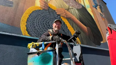 A man stands in the blue bucket of a cherry picker in front of a large, colourful wall painting. He is smiling and wearing a cap, dark hoody and black gloves. 