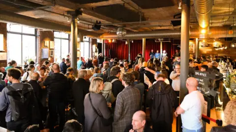 A crowd of people are seen chatting in an event space done out in an industrial style, with large, silver AC ducting on the ceiling. In the distance is a stage and a red curtain.