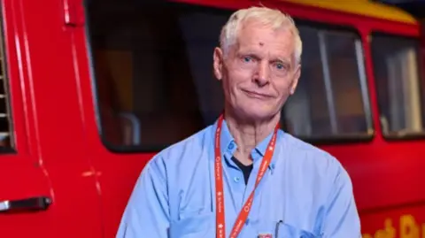 Royal Mail postman Robert Hudson looks towards the camera with a postvan in the background. robert is wearing a blue shirt with a red royal mail lanyard around his neck