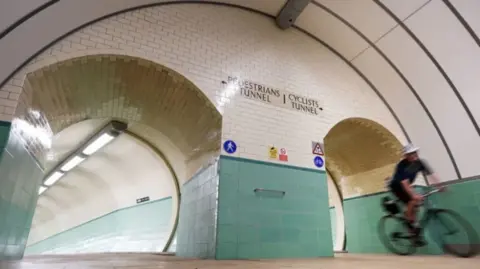 Two tunnels covered in green and cream tiles. There is a sign pointing to one which reads 'pedestrian tunnel' and one pointing to the other saying 'cyclists tunnel'. A man on a bike, dressed in black and a helmet is coming out of the cyclists tunnel and appears blurred.