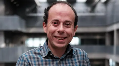 BBC A man with short brown hair and a grey and blue checked shirt smiles at the camera. He is standing in the atrium of a modern building, with the floors visible behind him.