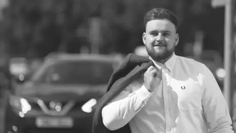 Family Handout Luke Allen in a black and white photo. He is wearing a shirt and carrying a jacket over his shoulder.