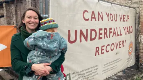 Sarah Murray, wearing a green coat and holding her baby, who is facing away from the camera. She is standing next to a banner which reads "can you help underfall yard recover?"