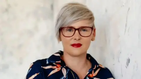 PEGS A woman with short blonde hair is wearing red-framed glasses and a blue shirt with red and white flowers on. She is looking at the camera and standing against a pale background. 