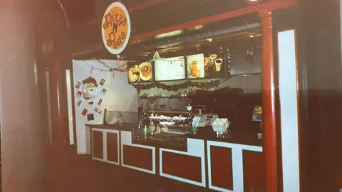 Philip Shelton A small food‑court stall with a sign above it that reads 'Pizza N Pasta'. The front counter has red and white paneling, and various items are displayed on the countertop.