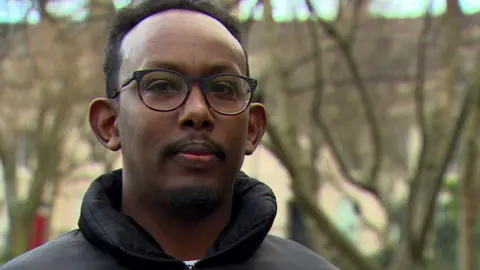 A man is looking into the camera. He's wearing glasses and a coat. He's standing outside. There are trees behind him.