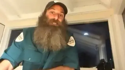 Sandy Duthie, hombre con barba grande y camisa verde 'Parks Victoria' mirando a la cámara, con el faro brillando en la oscuridad afuera, y un par de binoculares junto a él.