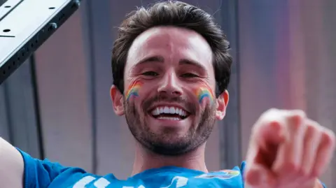 A Pride participant with rainbow face paint on both cheeks looks toward the camera while wearing a blue T-shirt and pointing forward. The image is photographed from a low angle against a backdrop of city buildings.