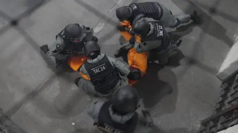 Ministry of Justice A view from above of five people, wearing black helmets, black stab vests and grey uniforms appearing to roll up orange fabric in a prison space with a dark grey concrete floor.