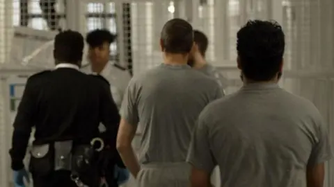 BBC Men wearing prison clothing are escorted in a line by a uniformed prison officer inside a prison in north London