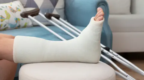 Getty Images A leg in a cast is seen lying across a bed with cutches propped in the background