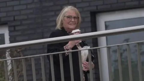 Sharman behind a metal silver fence walking into court. She is smiling and looking at the camera. She carries a takeaway coffee and a bag in her other hand.