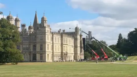 Stately home - Burghley House with a big lawn in front of it and red and green cranes parked next to the building.